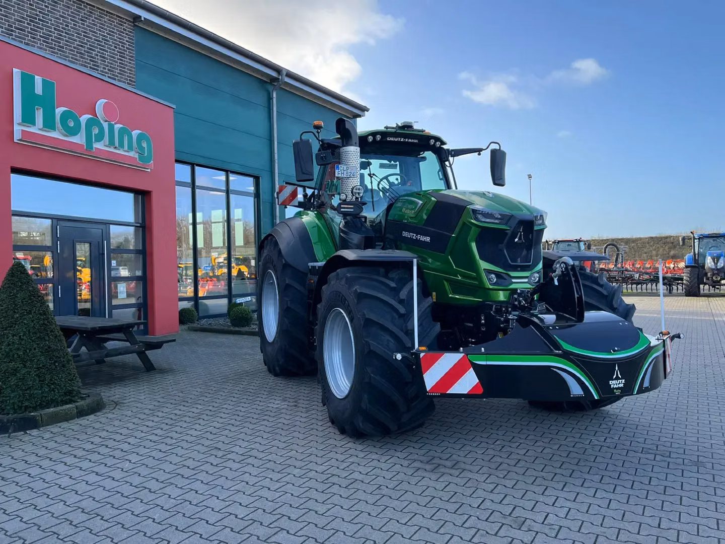 A green tractor with a front weight bumper attached is parked on paving stones outside a building with large windows.