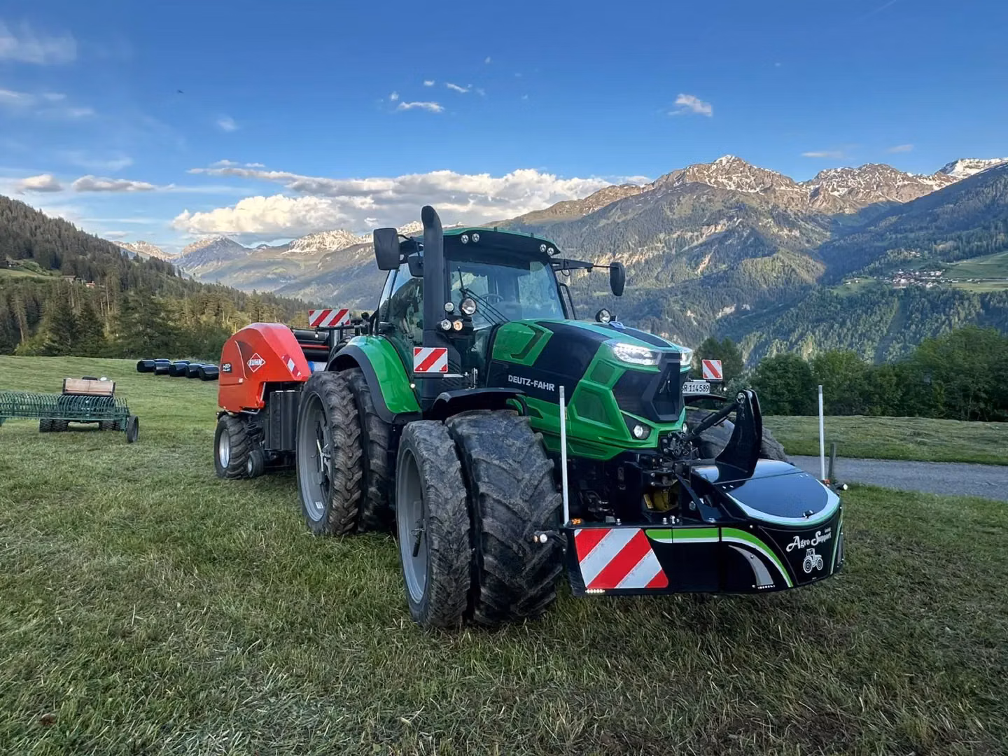 Green tractor with a front weight bumper attaches to a red agricultural implement on grass with mountains in the background