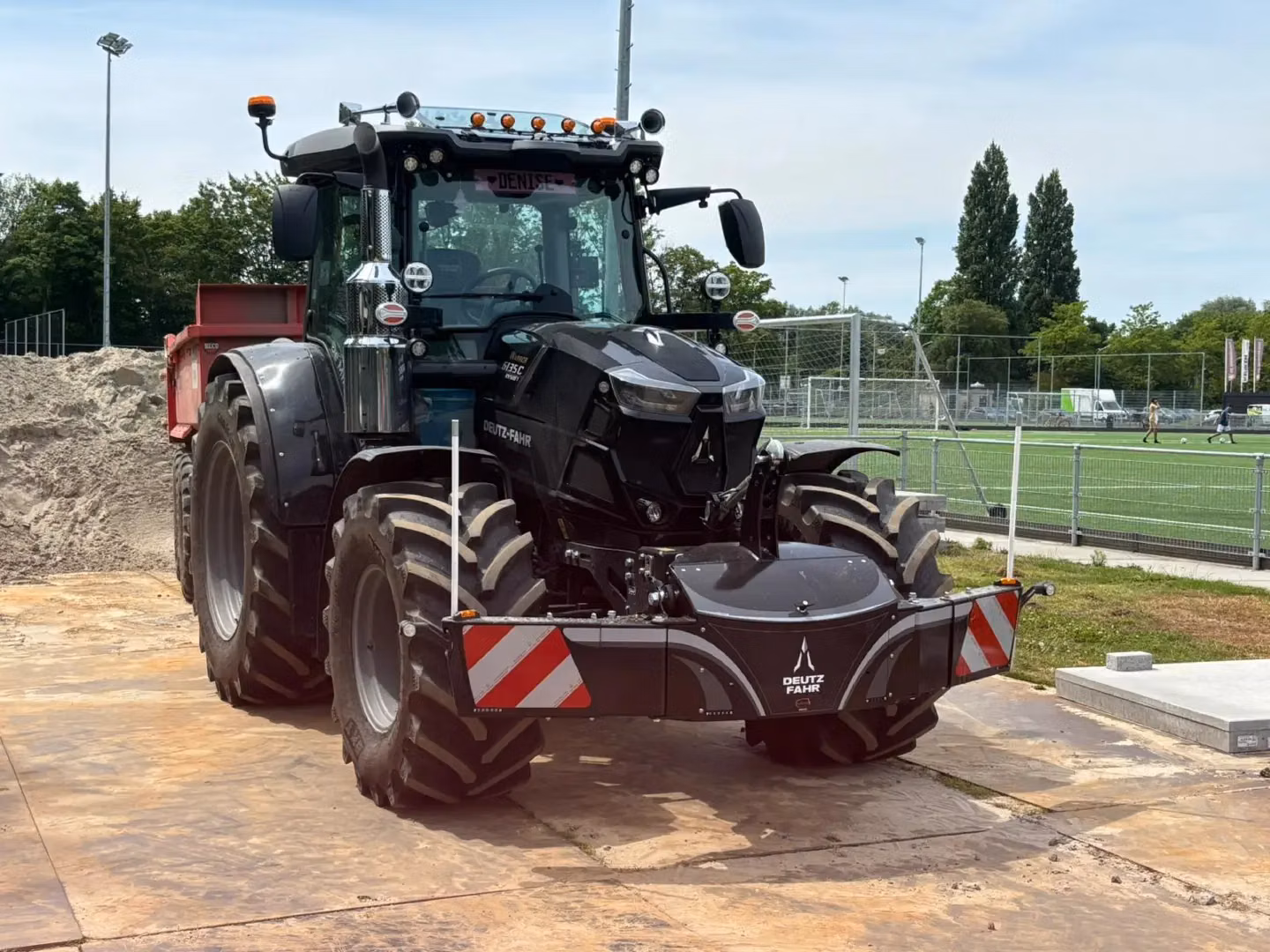 A black tractor with a red trailer and a Deutz-Fahr front weight bumper is parked on a construction site near a sports field