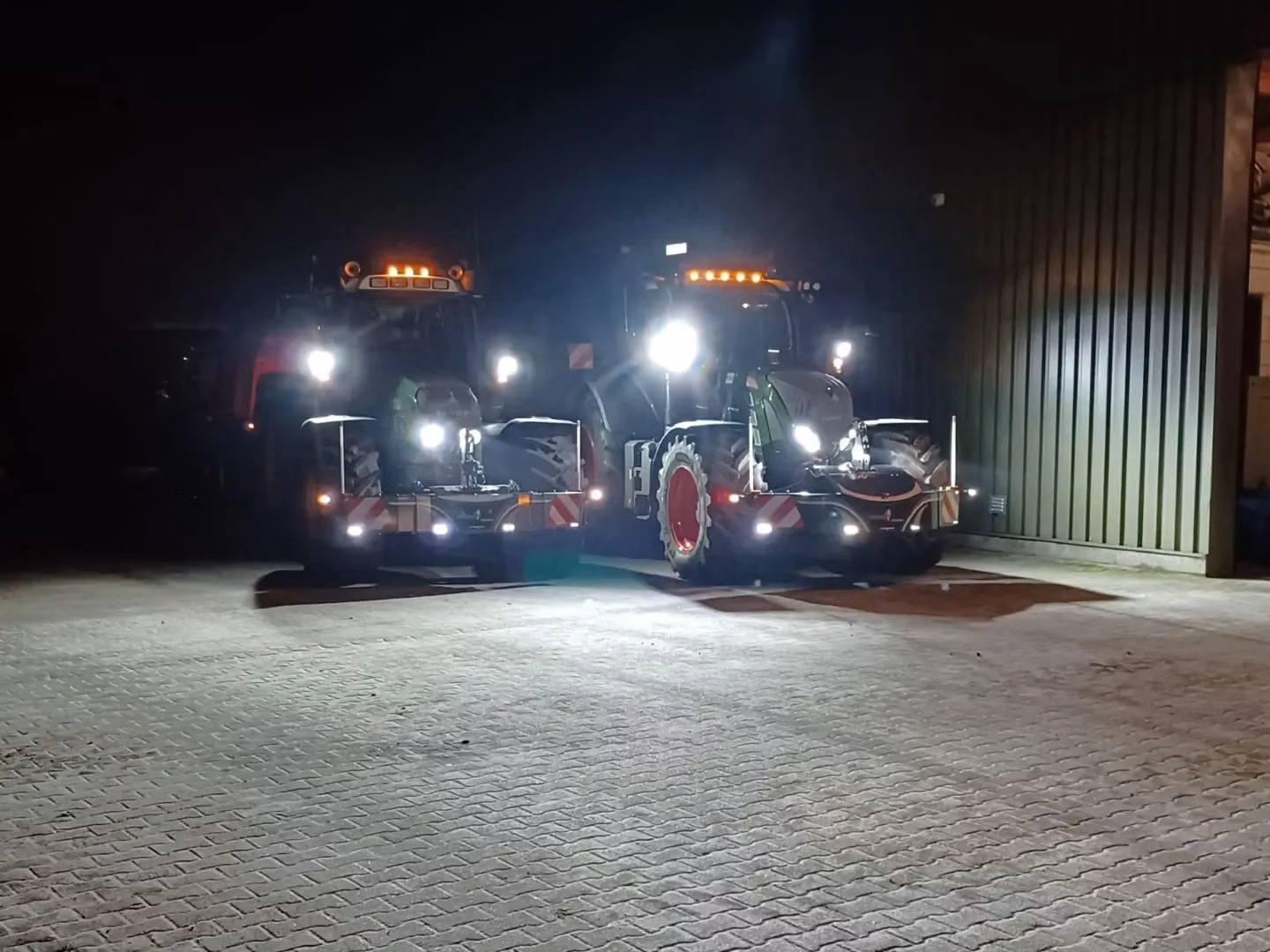 Two tractors with front weight bumpers and headlights on are parked outside a building at night
