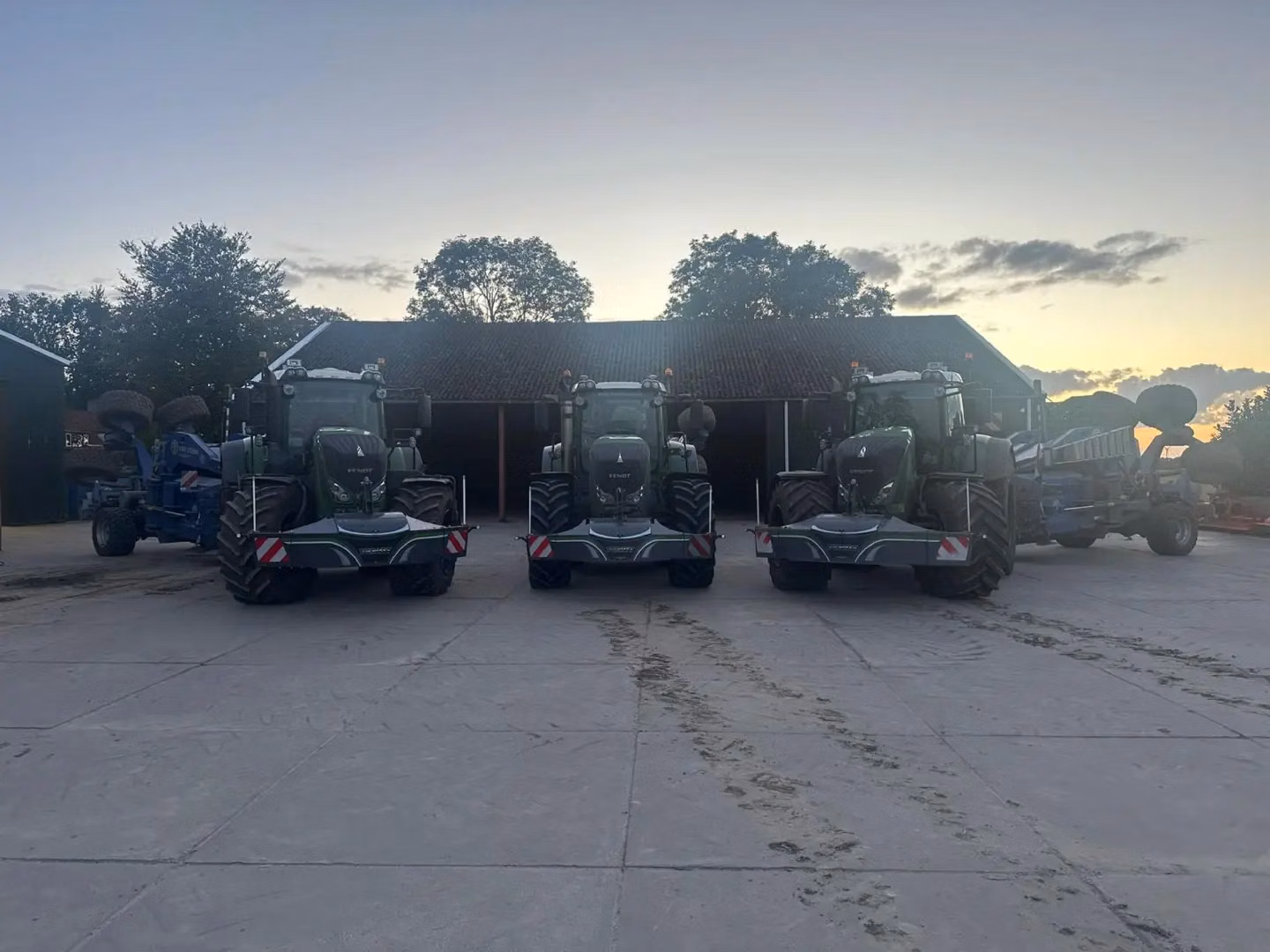 Four tractors with front weight bumpers are parked side by side in front of a barn at sunset