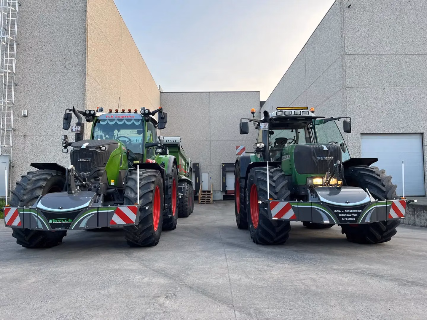 Two green tractors with front weight bumpers and red wheels parked on concrete between industrial buildings