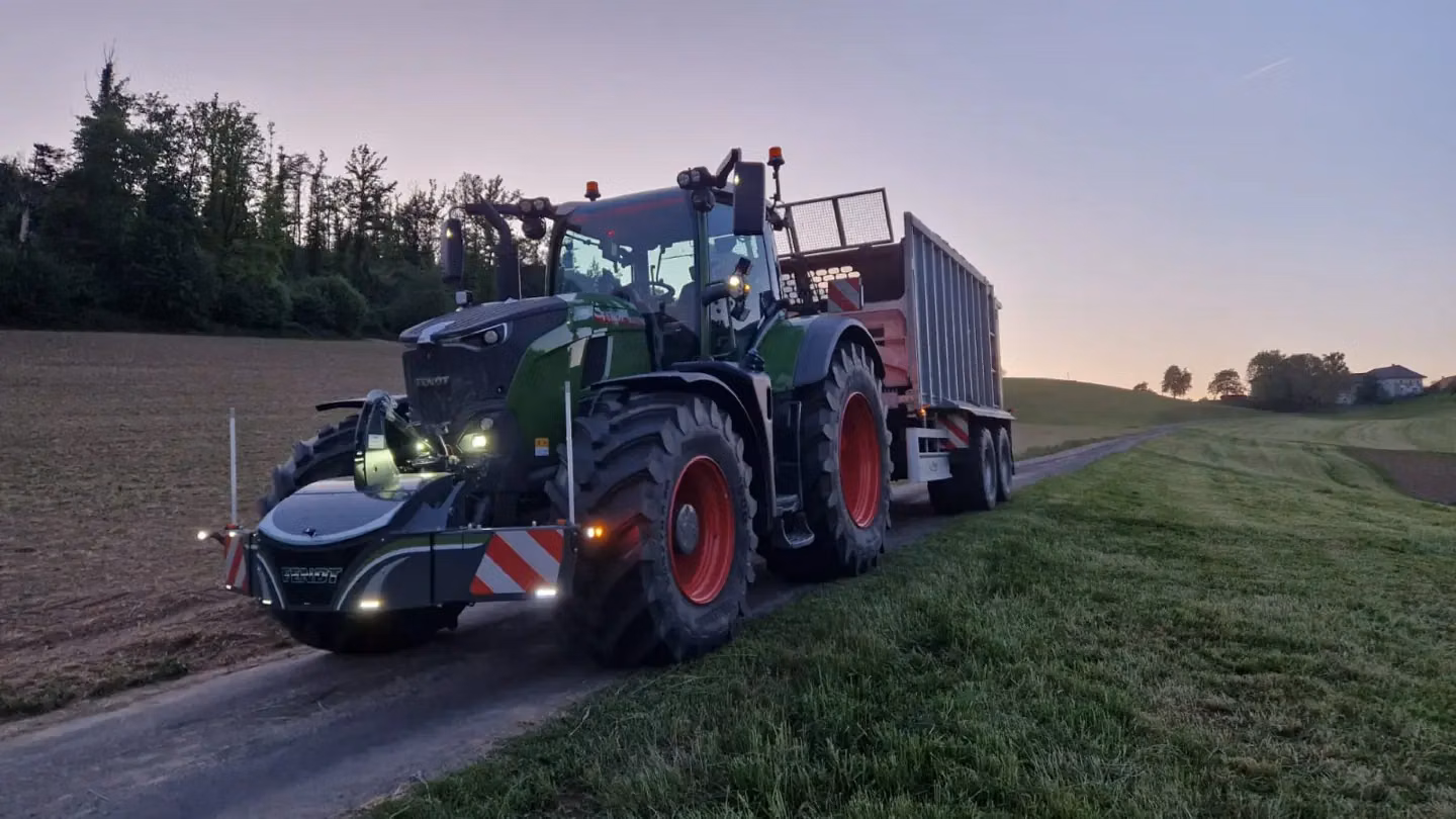 A green tractor with a front weight bumper and attached trailer stands on a rural road at dusk
