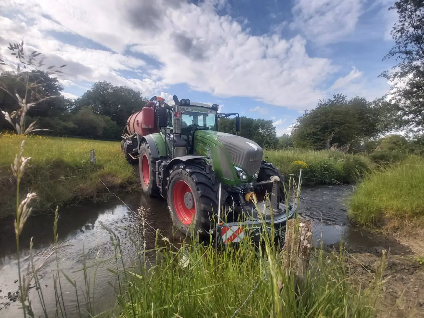 Green tractor with front weight bumper crossing a shallow stream in a grassy rural landscape under a partly cloudy sky