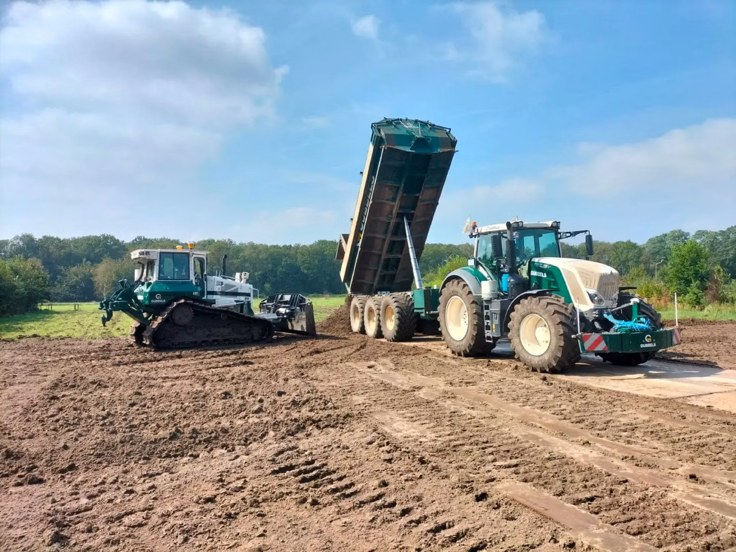 A green tractor with a front weight bumper and trailer, next to a tracked bulldozer on a muddy field