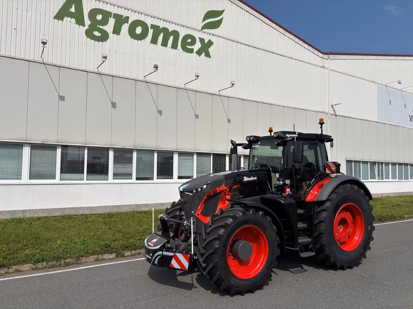 Black tractor with red wheels parked on a road, fitted with a front weight bumper, in front of a building marked Agromex