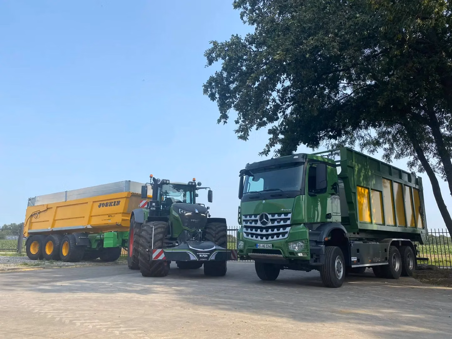 A tractor with a front weight bumper and yellow trailer parked beside a green and yellow truck under a tree