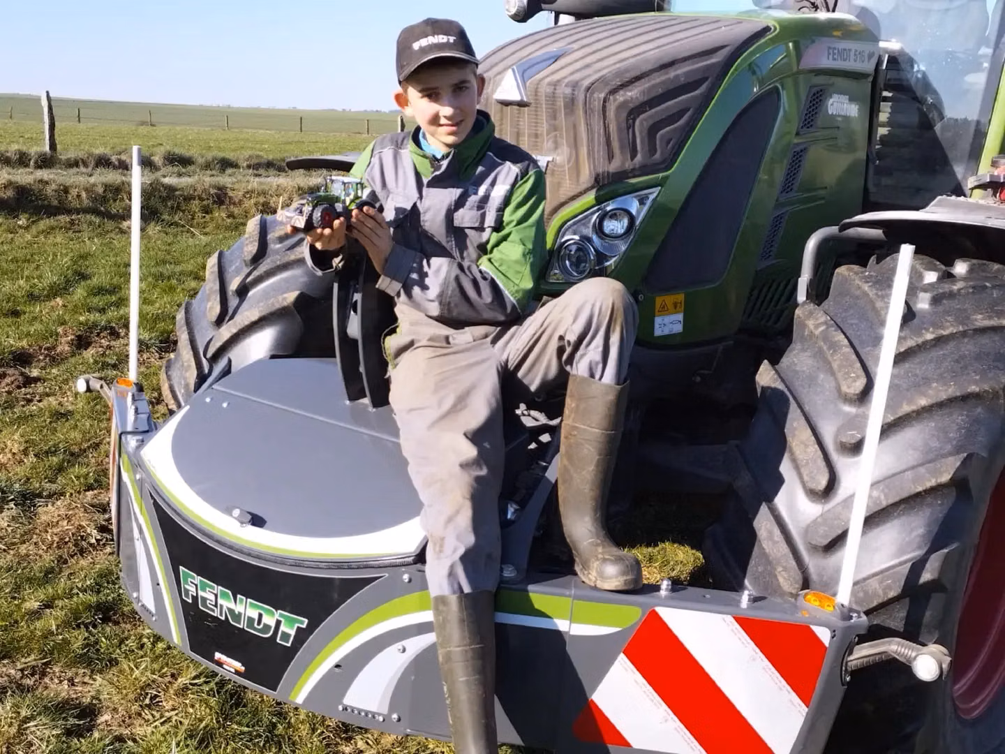 A person in work clothes sits on a Fendt front weight bumper attached to a tractor, holding a toy tractor.