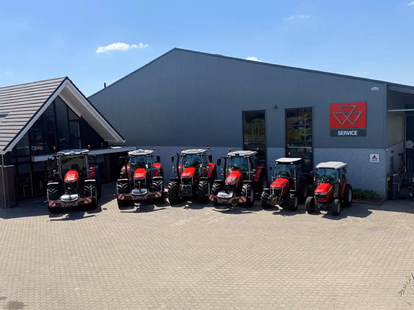 Seven red tractors with front weight bumpers are parked in front of a building with a service sign