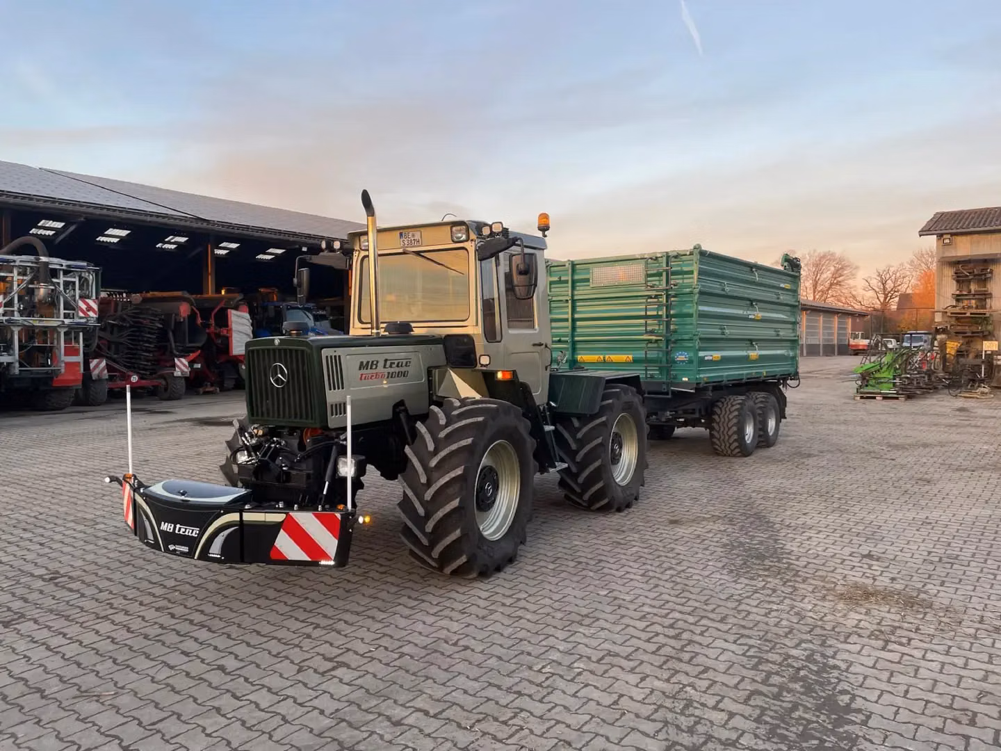A tractor with a front weight bumper and attached trailer is parked on a paved farmyard near equipment sheds