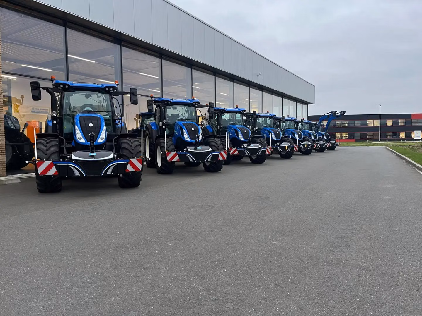 Seven blue tractors with front weight bumpers are lined up outside a building on a paved lot