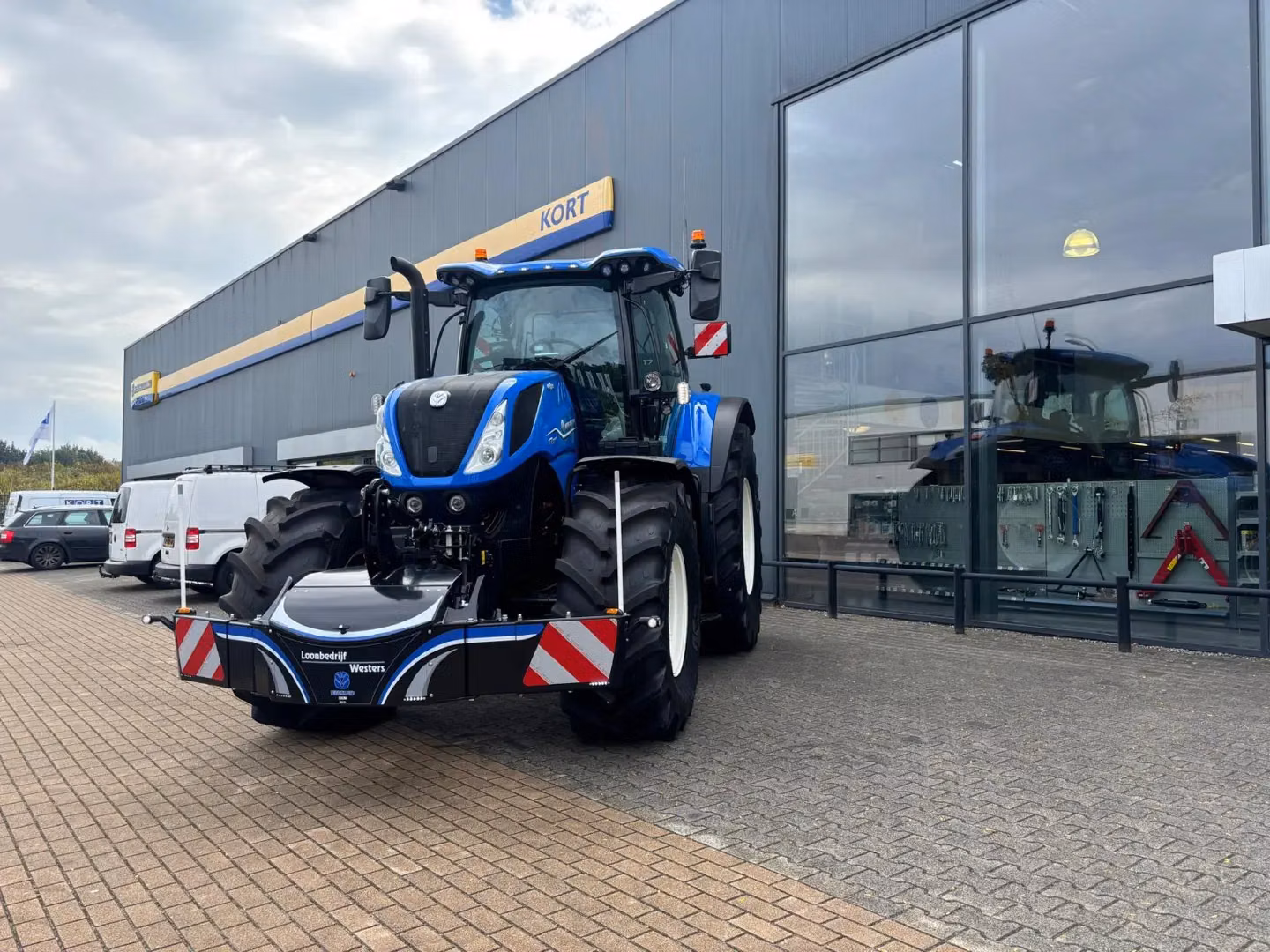 Blue tractor with a front weight bumper parked outside a building with large windows and several vehicles nearby