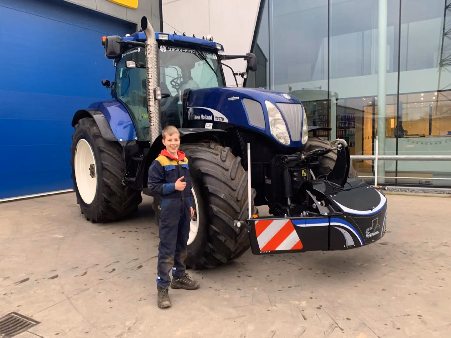 A boy in work clothes stands smiling in front of a blue tractor fitted with a front weight bumper box