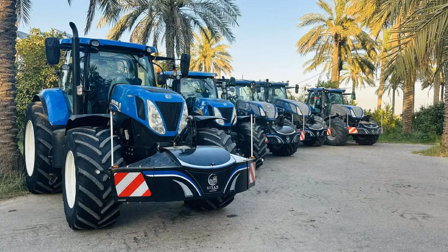 Five blue tractors with front weight bumpers parked on a road lined with palm trees