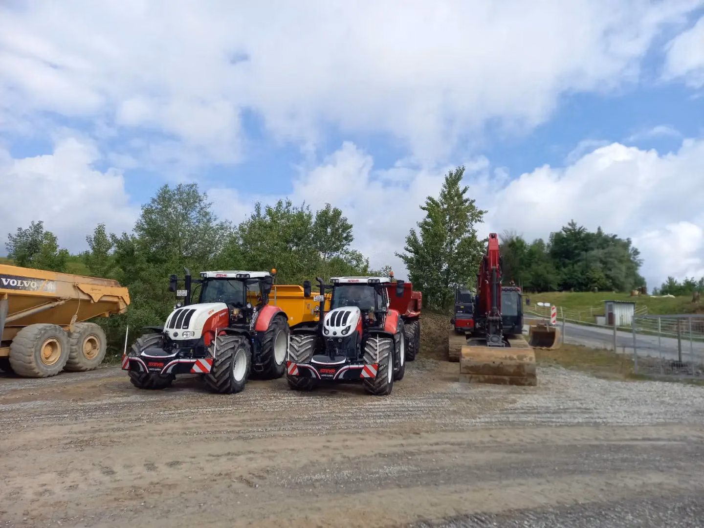 Two tractors with front weight bumpers are parked on gravel beside construction vehicles and trees under a partly cloudy sky