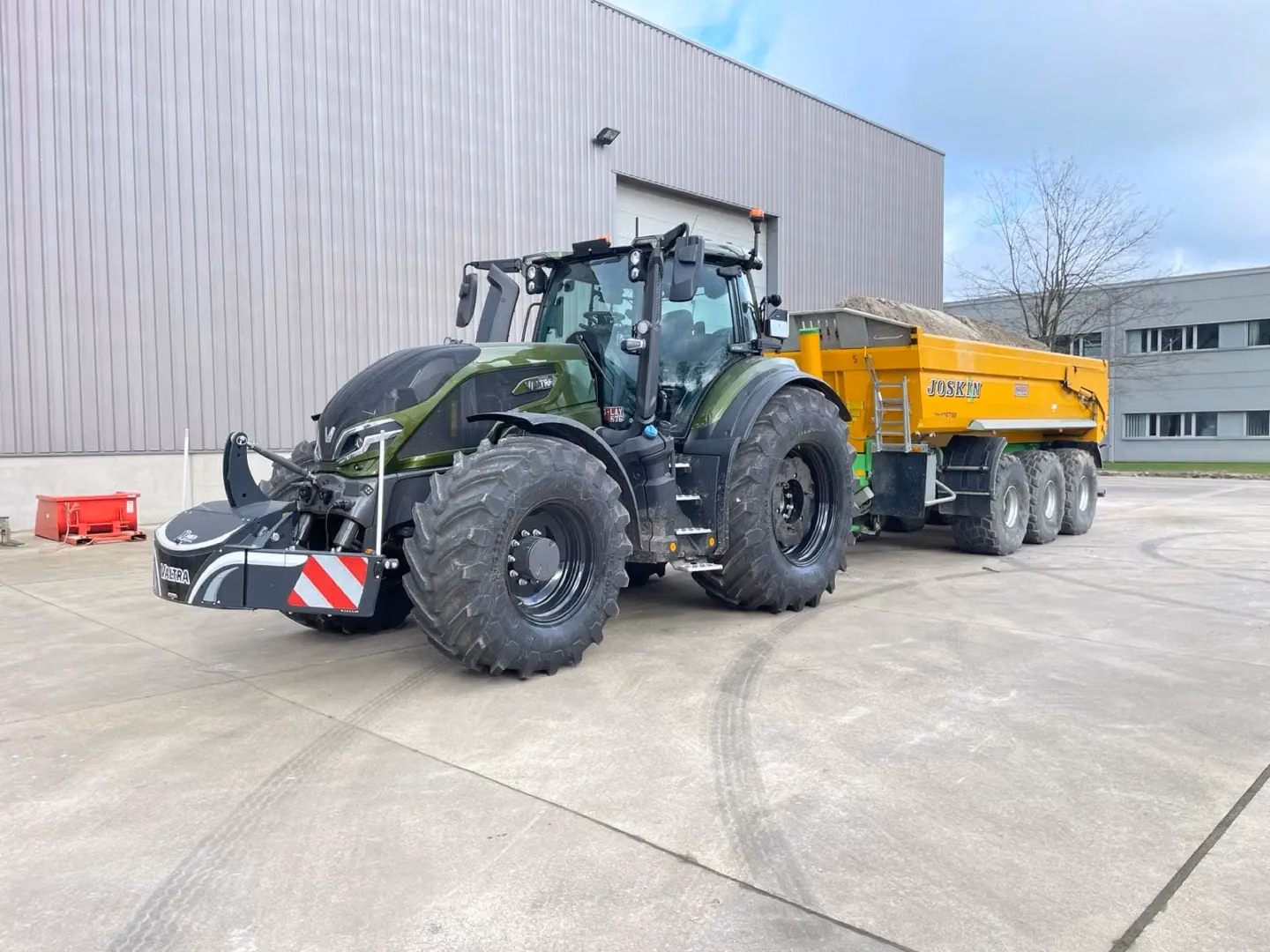 A green tractor with a front weight bumper featuring LED lights is attached to a yellow trailer in a yard