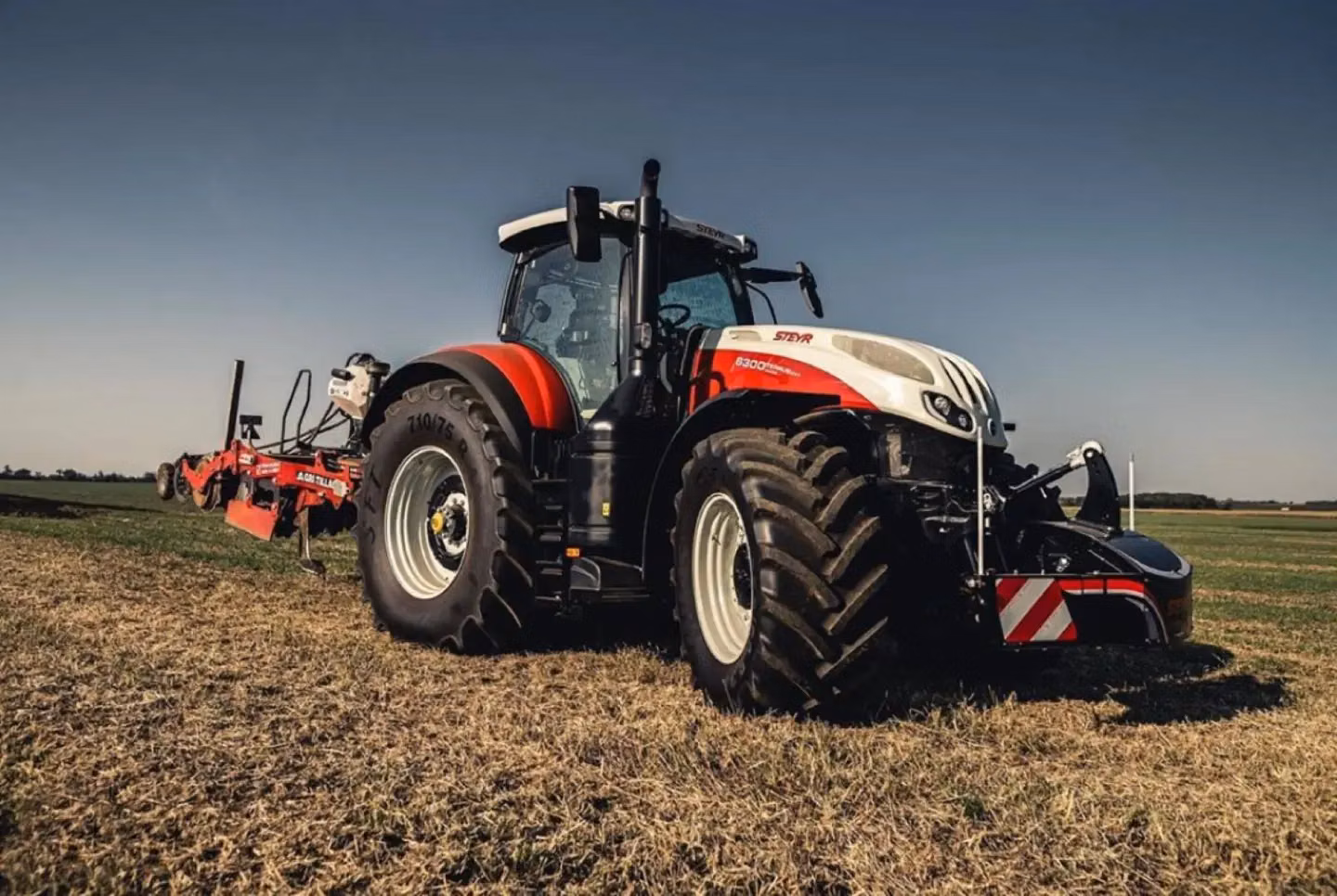 A red and white tractor with a front weight bumper attached, parked on a grassy field under a clear sky