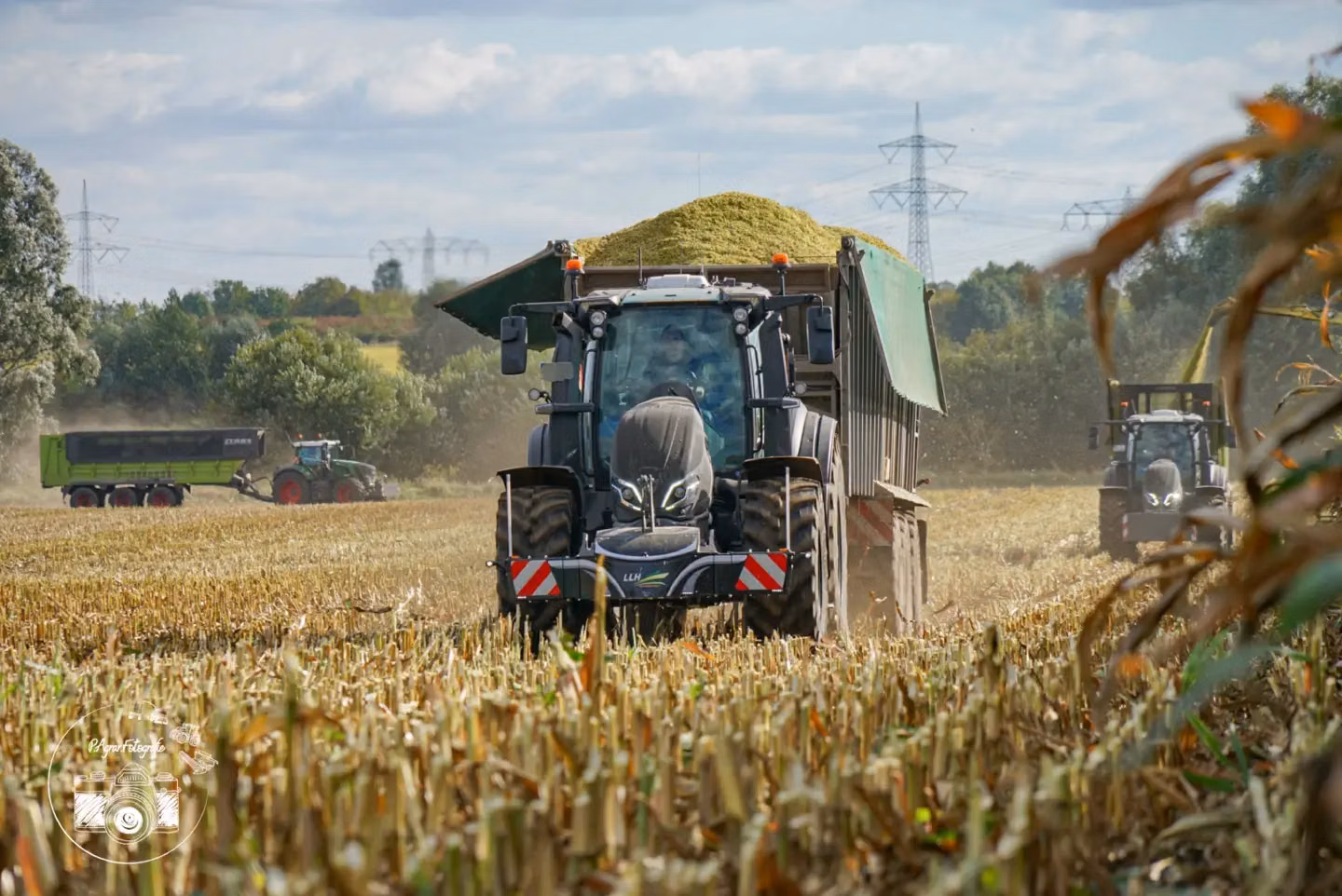 Black tractor with a front weight bumper towing a loaded trailer on a harvested field, two more tractors in background