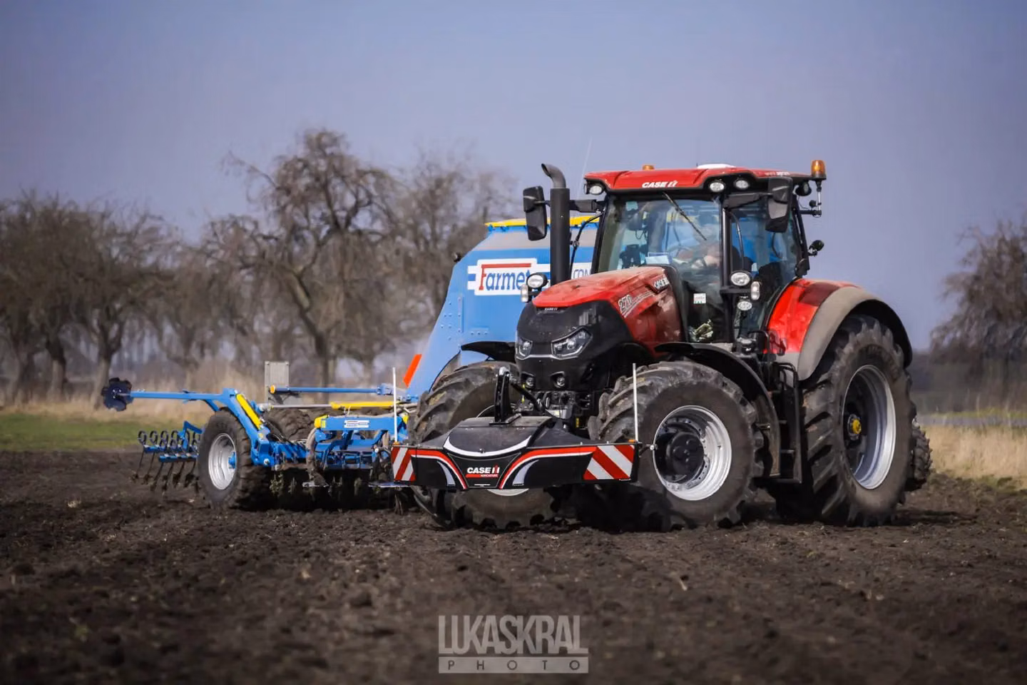 A red tractor with a front weight bumper attached works in a field towing blue agricultural equipment