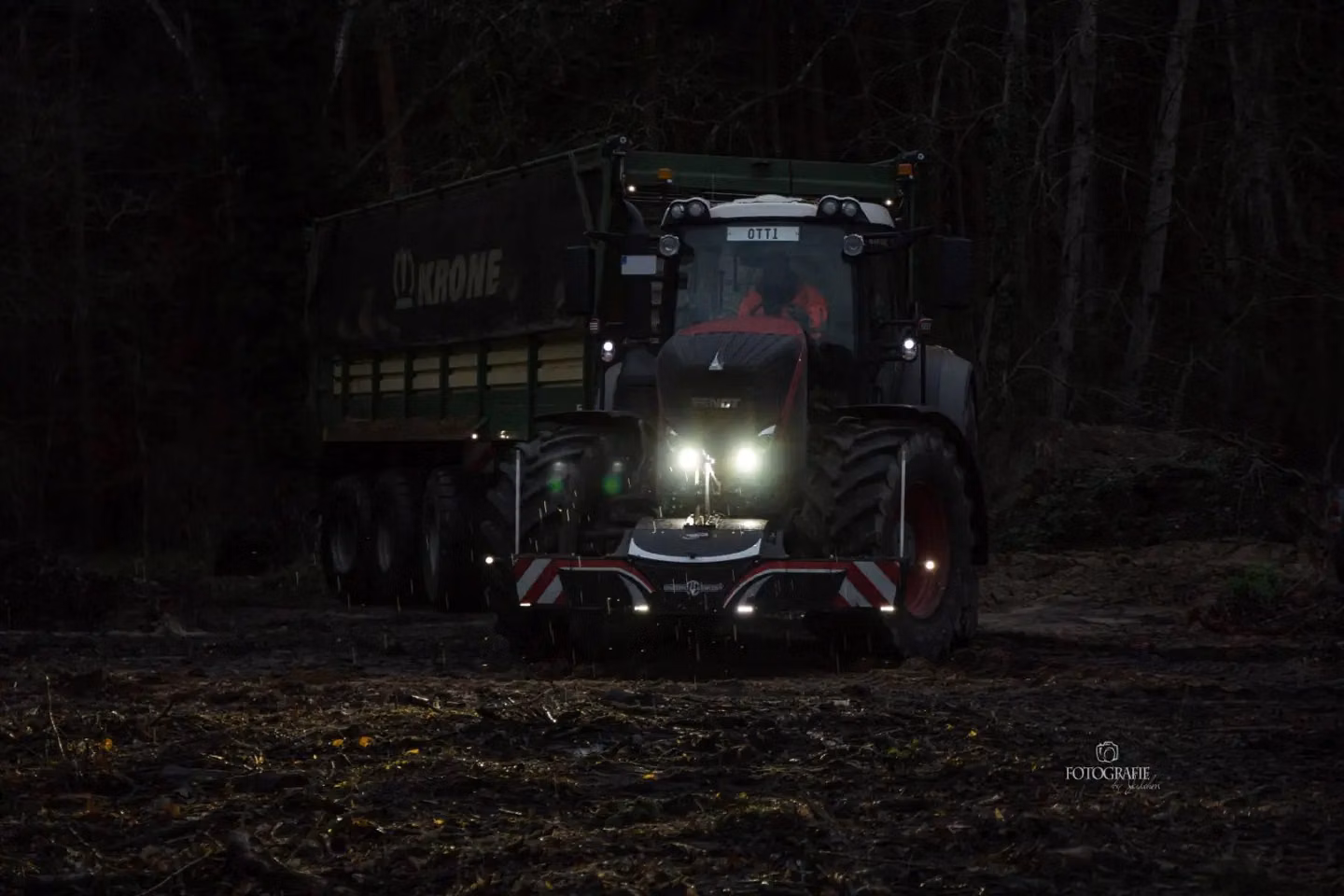 Tractor with front weight bumper featuring LED lights working at dusk in a forest clearing, trailer attached behind