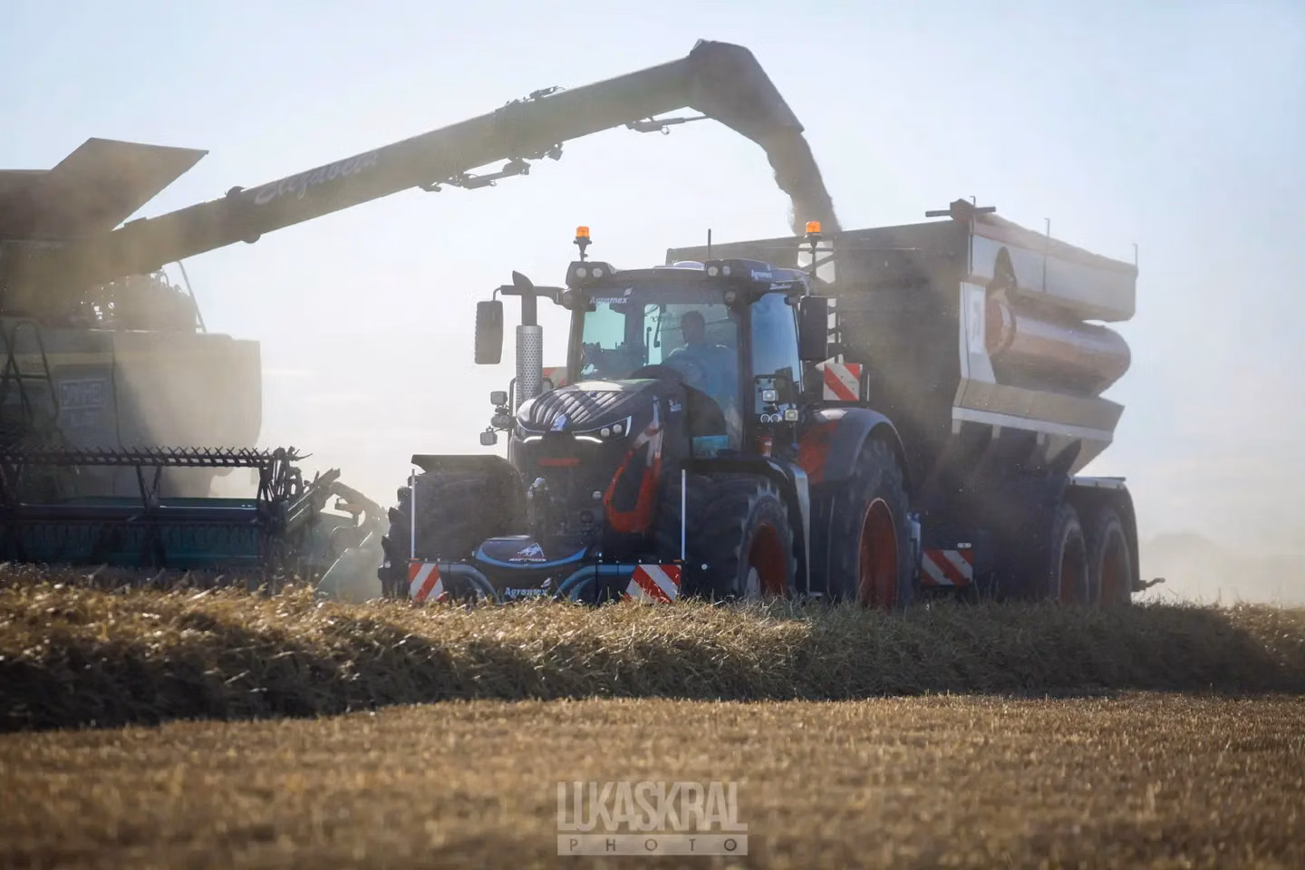 A tractor with a front weight bumper and lights is collecting grain from a combine harvester in a dusty field