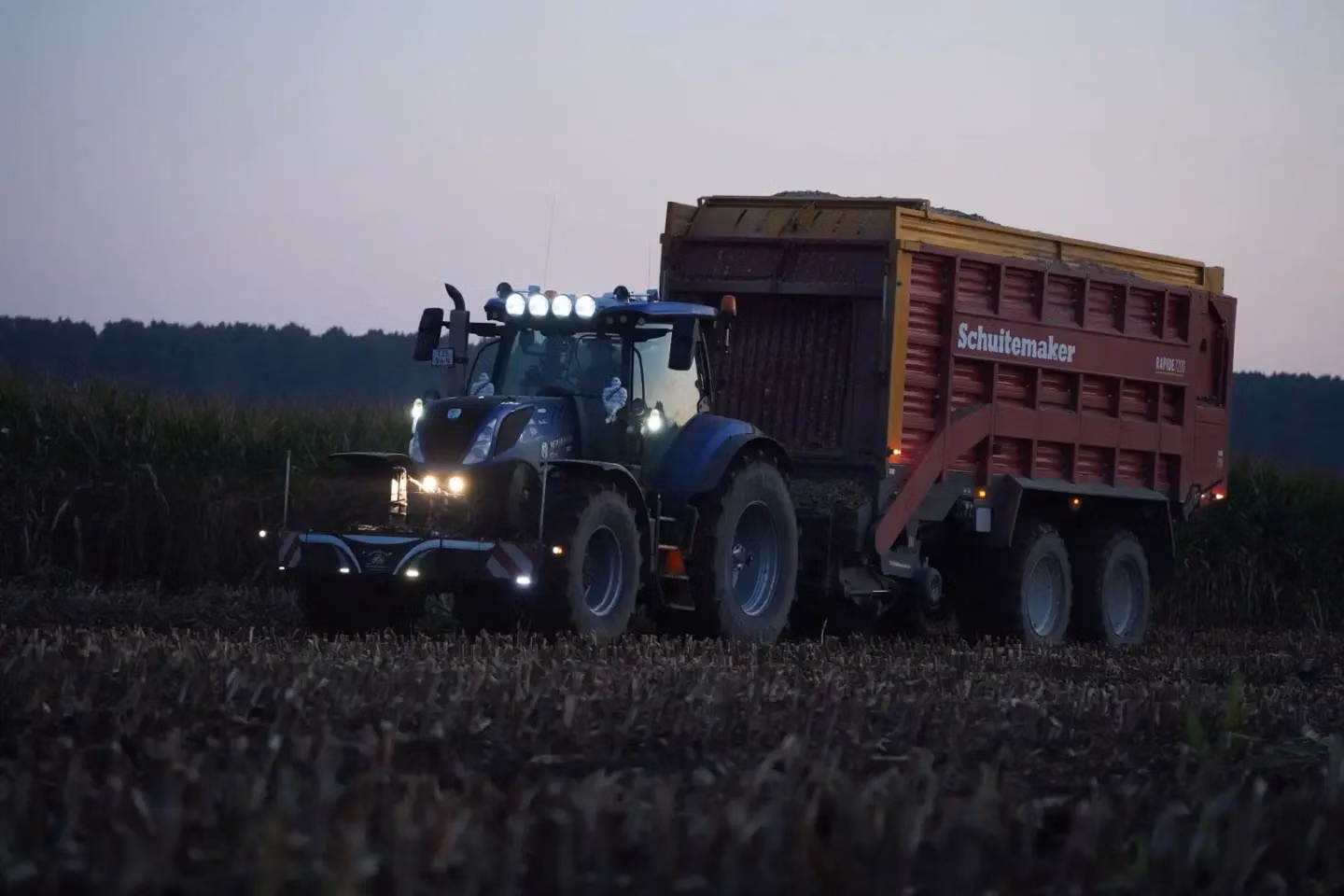 A blue tractor with an illuminated front weight bumper and large trailer drives through a harvested field at dusk