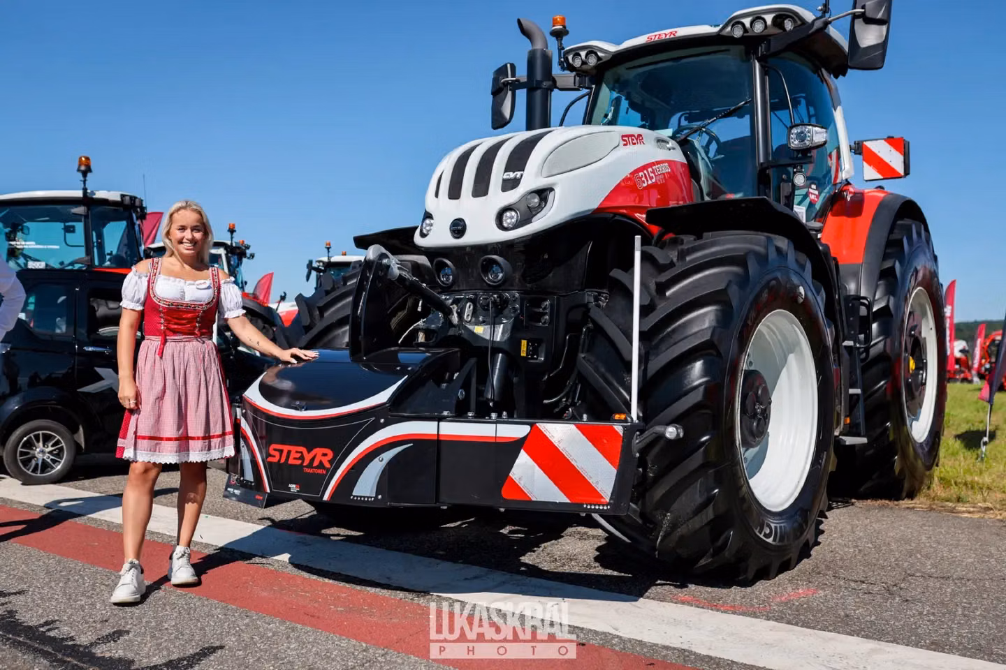 A woman in a dirndl stands beside a large tractor equipped with a Steyr front weight bumper on display outdoors