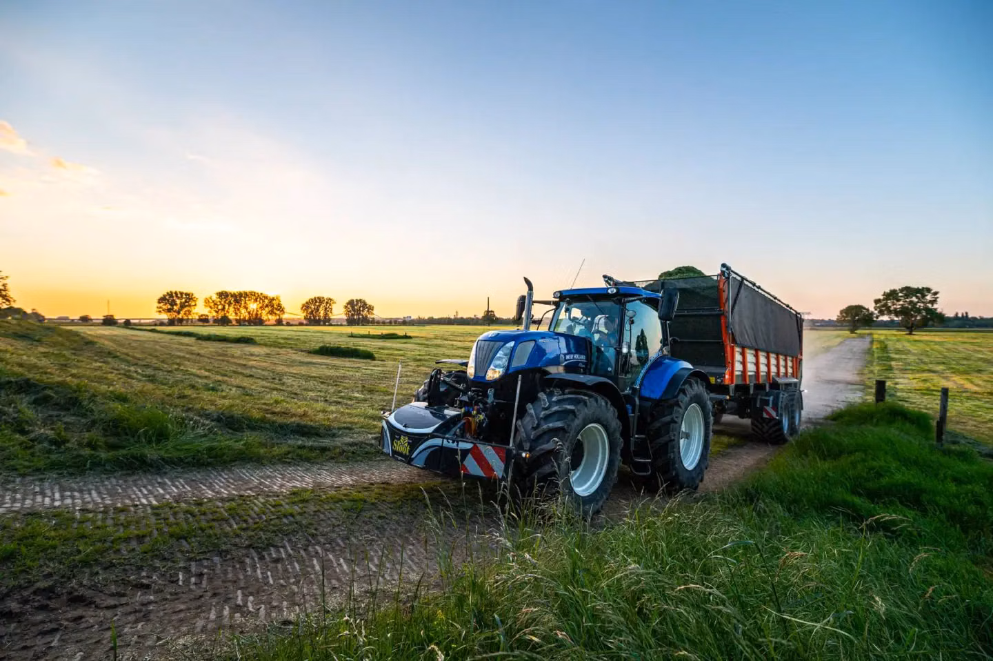 Blue tractor with a front weight bumper and trailer on a rural path at sunset with fields in the background