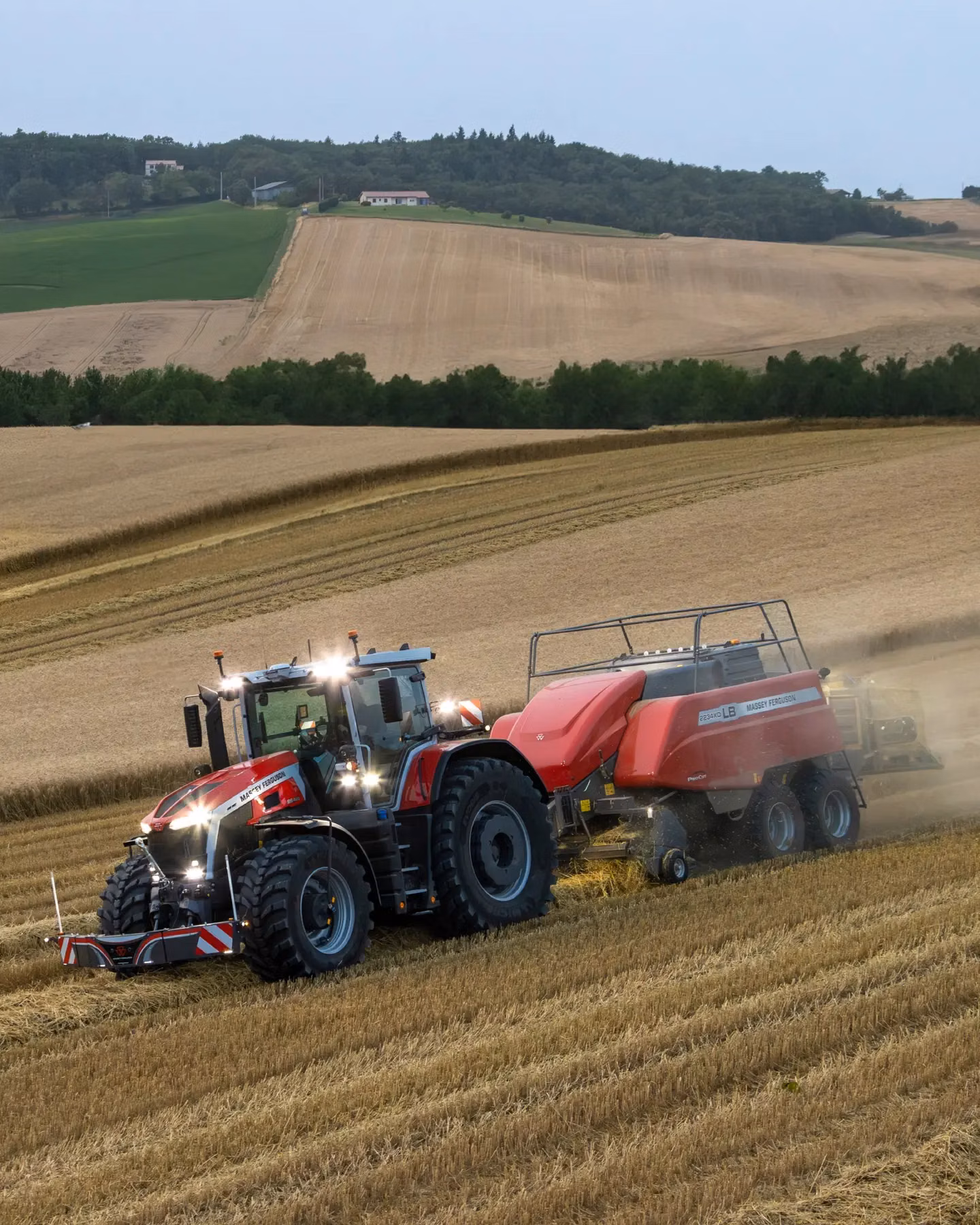 A red tractor with front weight bumper is pulling a large baler through a harvested field with hills in the background