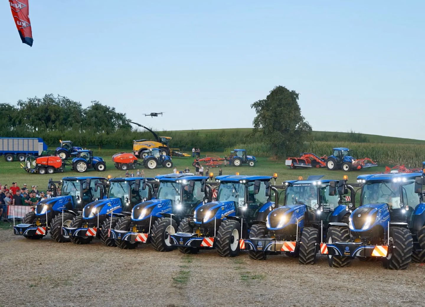 A row of blue tractors each fitted with front weight bumpers, with people and more machinery in the background