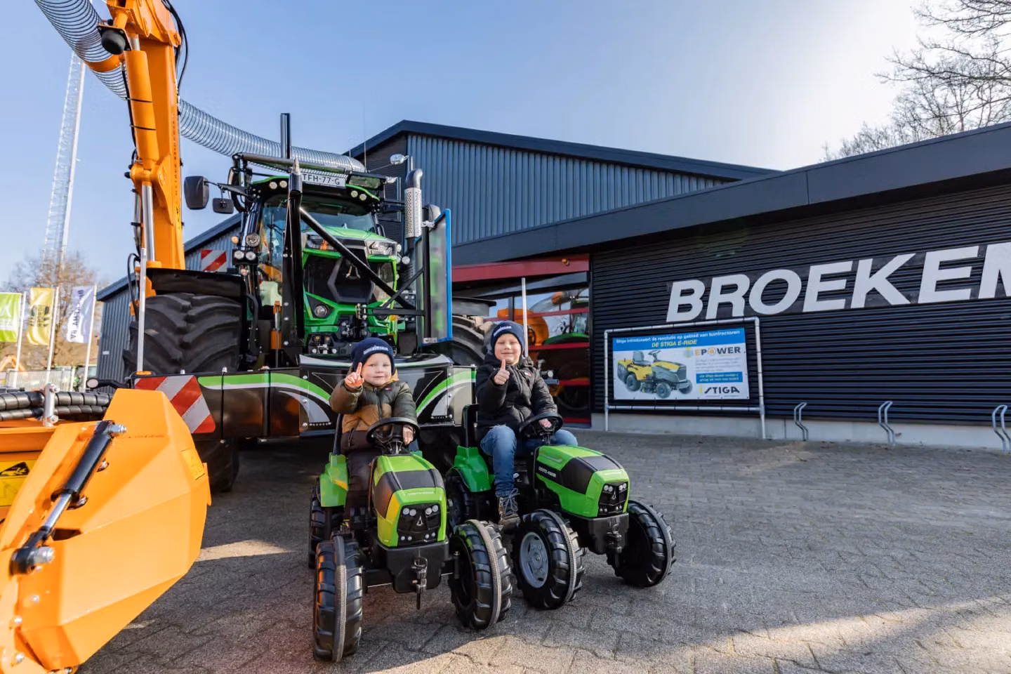 Two children on toy tractors are in front of a large tractor with a front weight bumper and orange attachment