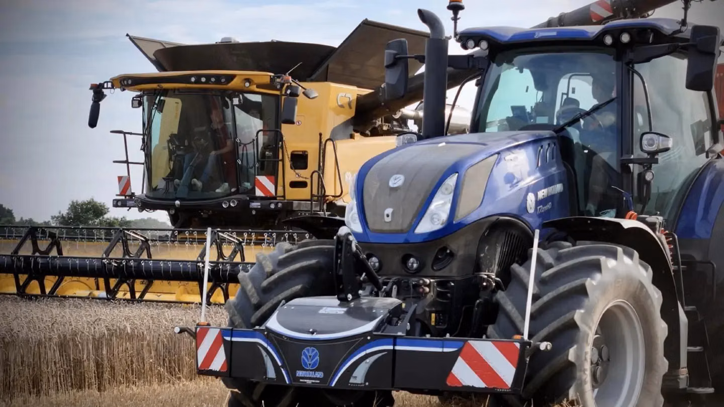 A blue tractor with a front weight bumper is parked in a field beside a large yellow combine harvester