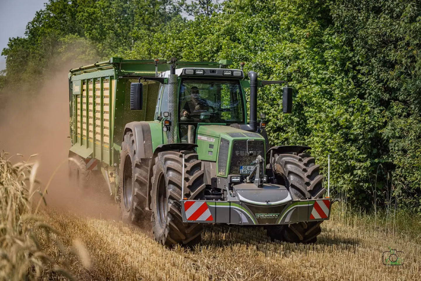 Large green tractor with a front weight bumper and lights drives through a dusty field next to tall green hedges