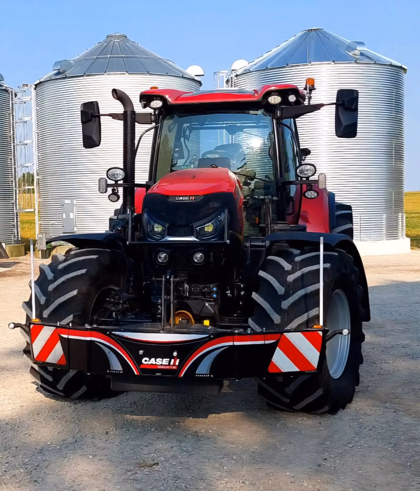 Red tractor fitted with a CASE IH front weight bumper, parked on gravel in front of silver grain silos