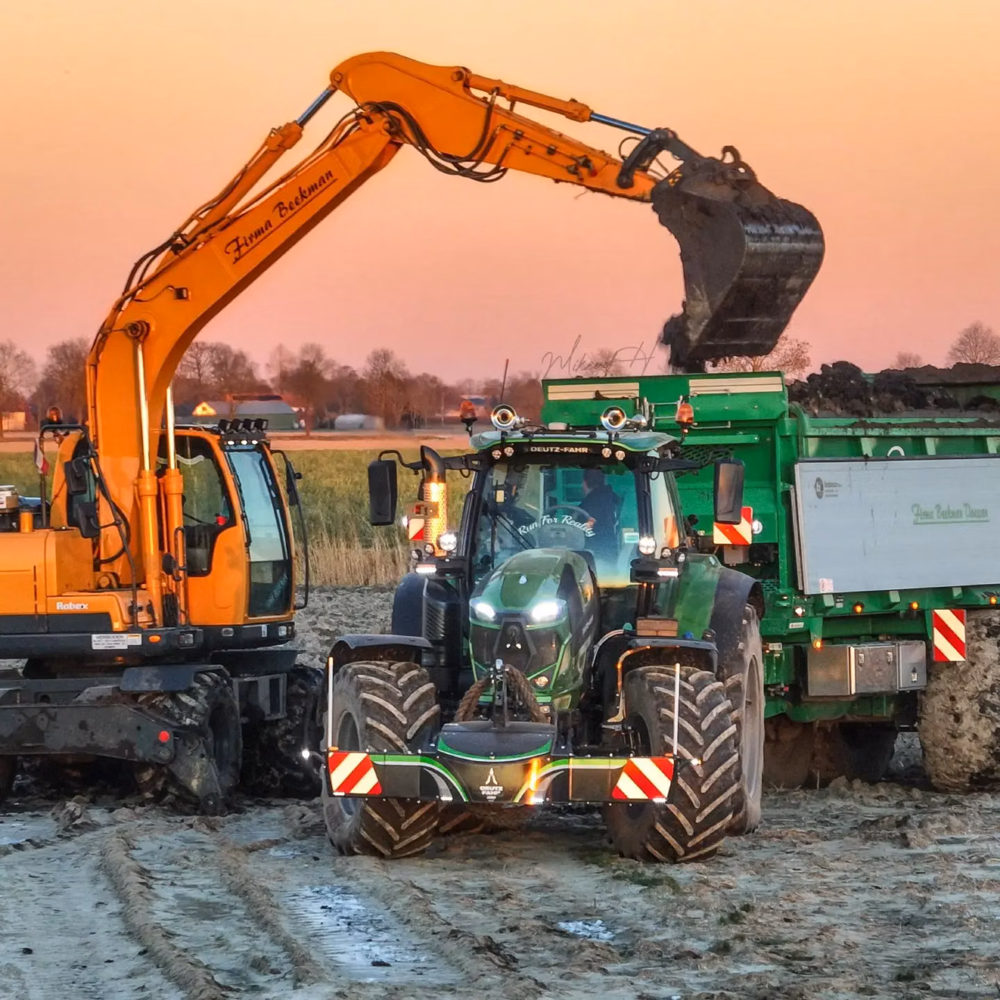 A green tractor with a front weight bumper is attached to a trailer while an excavator loads soil into it