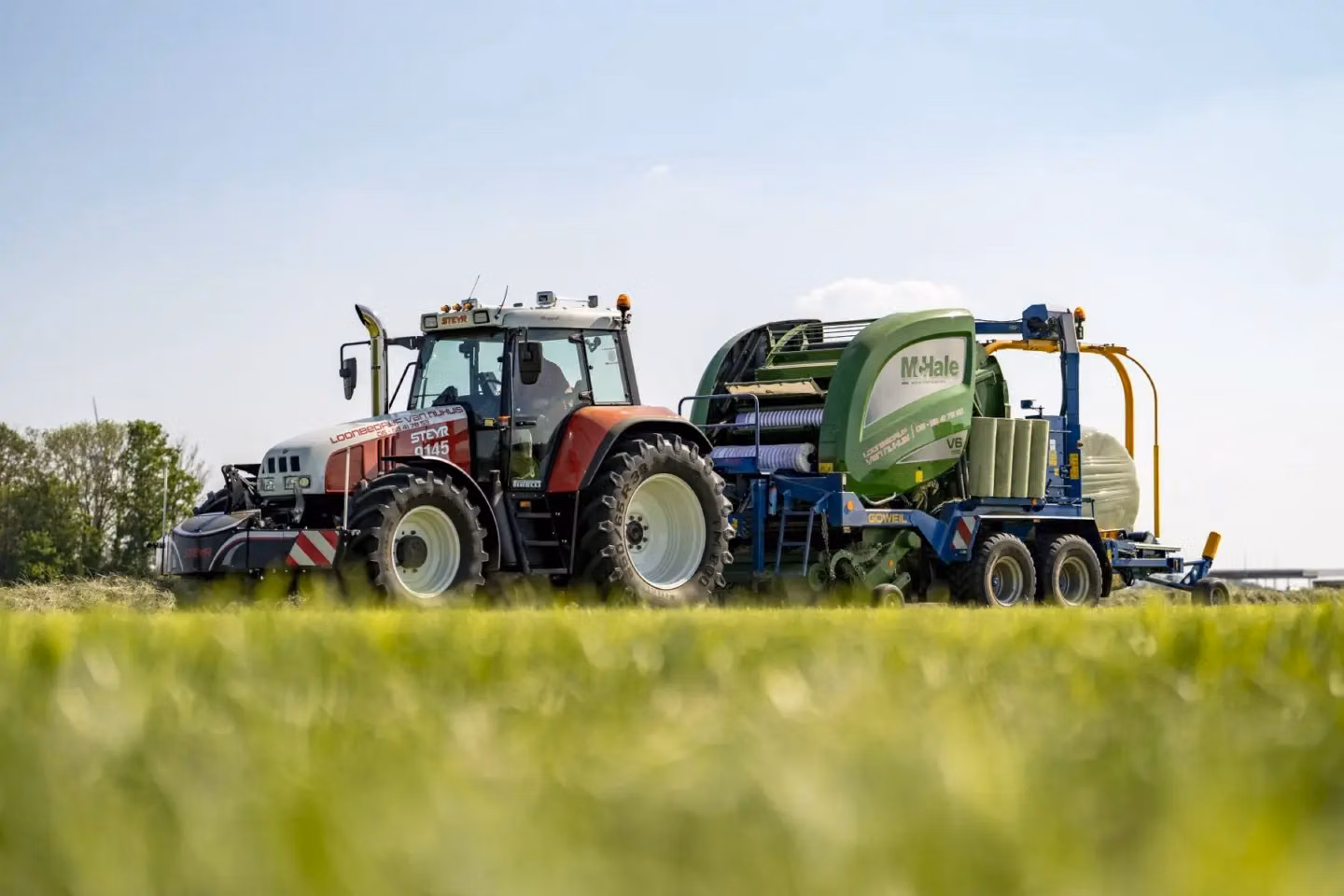 Red and white tractor with a front weight bumper attached, working in a grassy field with a green baler implement