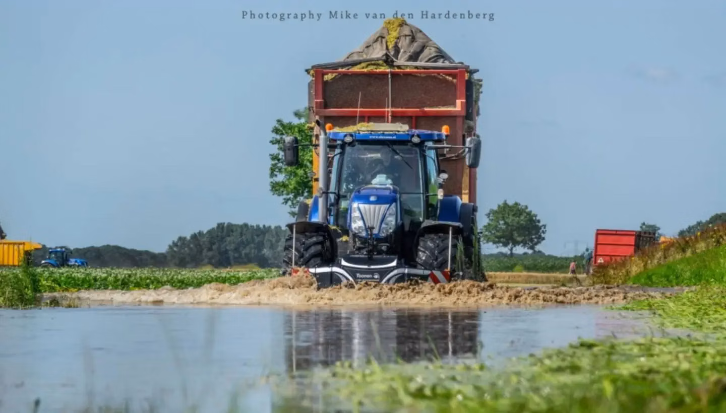 Blue tractor with a front weight bumper is driving through a flooded field towing a large trailer of harvested crops