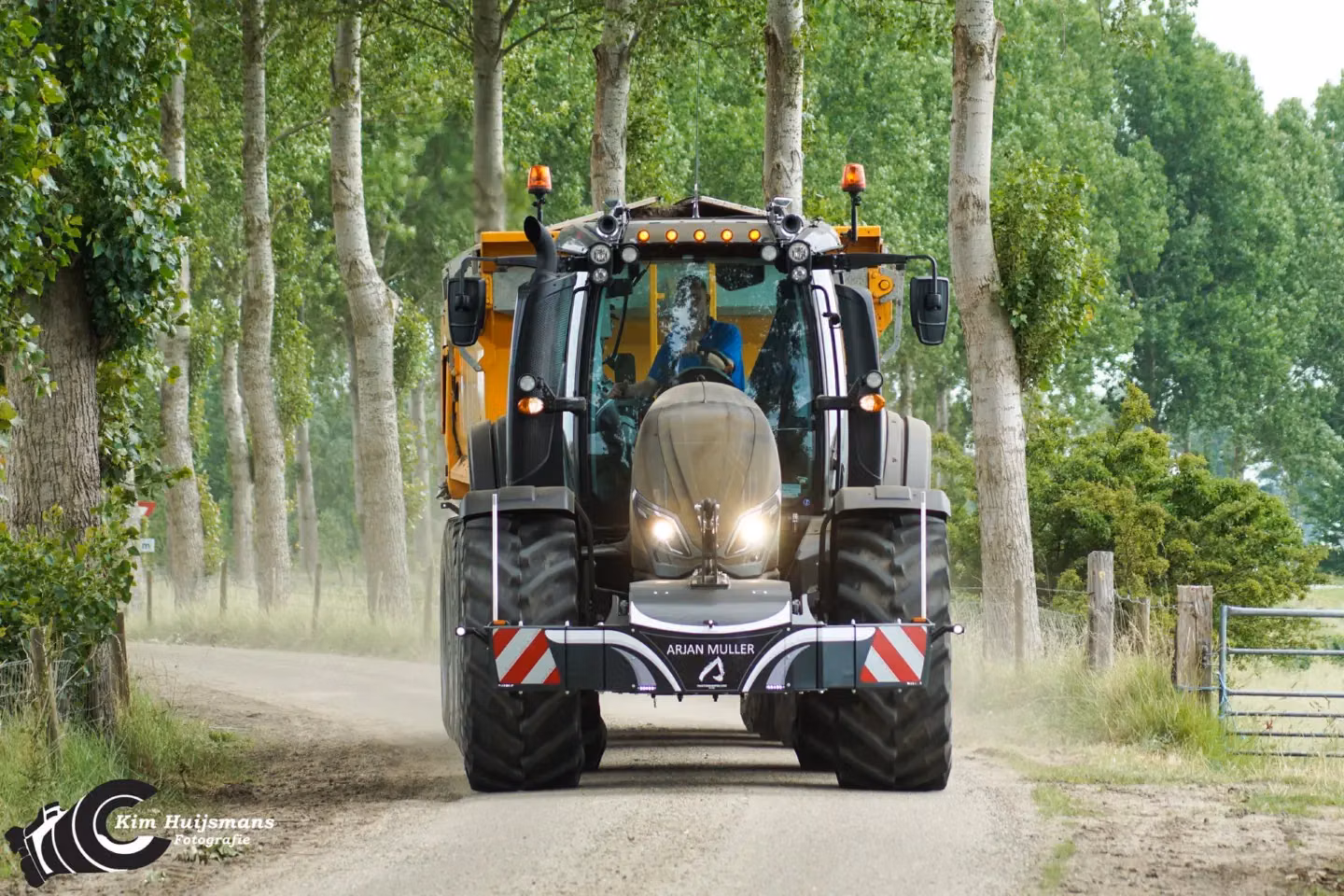 A tractor with an attached front weight bumper labeled "Arjan Muller" drives on a rural dirt road lined with trees