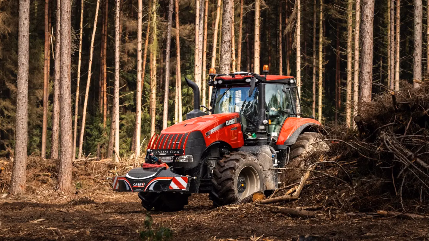 Red tractor with a front weight bumper is parked in a forest clearing beside a pile of branches and logs
