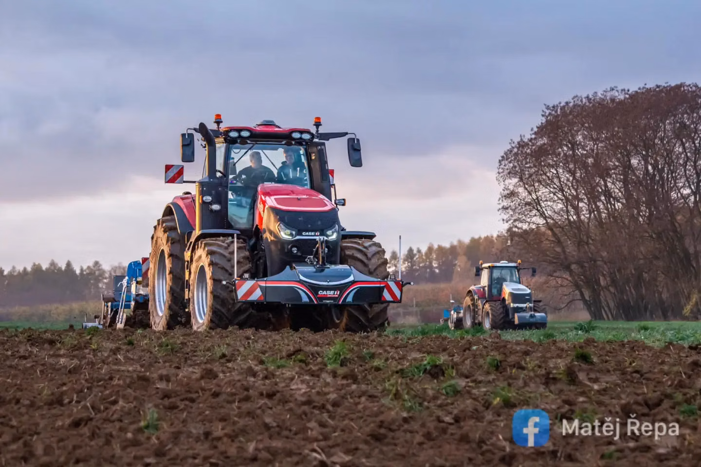 Red tractor with a front weight bumper working in a field, another tractor in the background, cloudy sky above