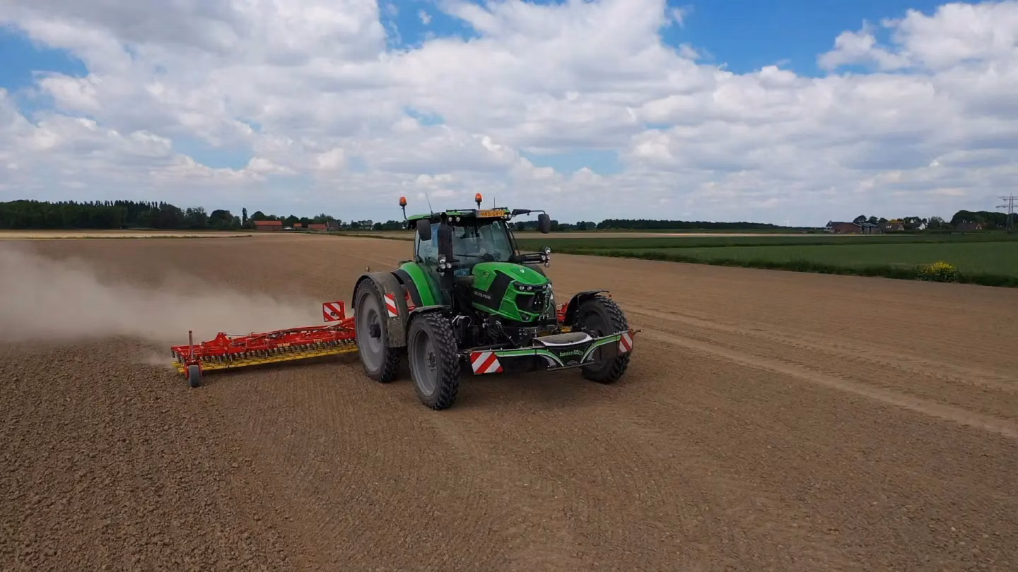 Green tractor with a front weight bumper and dust box attachment working on a plowed field under a cloudy sky