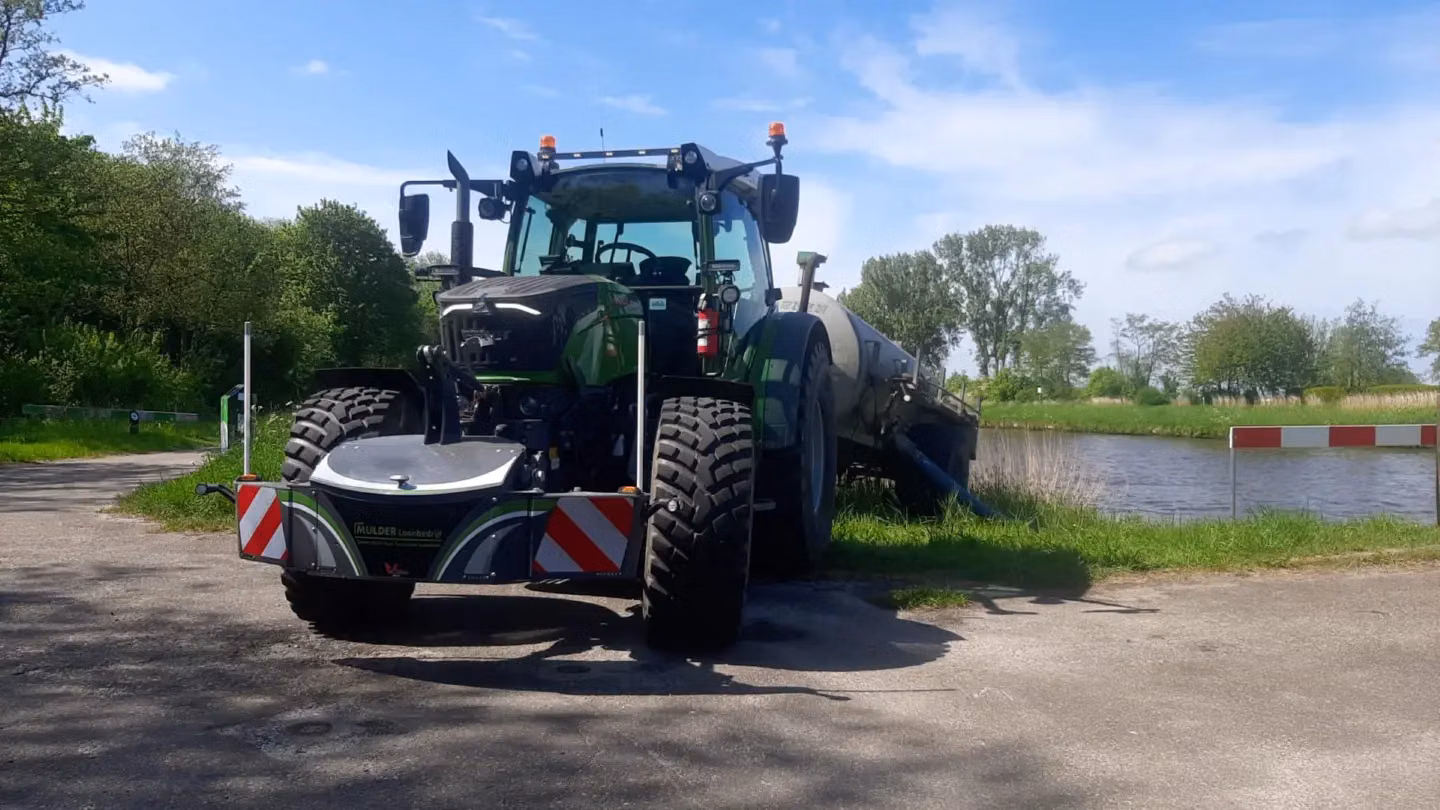 A tractor with a front weight bumper featuring reflective stripes is parked beside a waterway on a sunny day