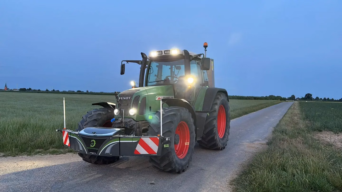 Green tractor with illuminated lights and a front weight bumper on a rural road at dusk