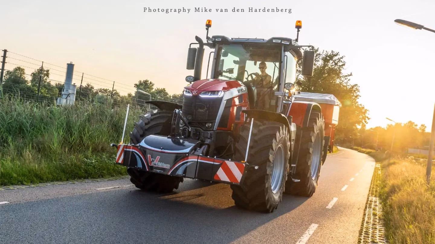 Red tractor with a front weight bumper and visible lights driving on a paved country road at sunset