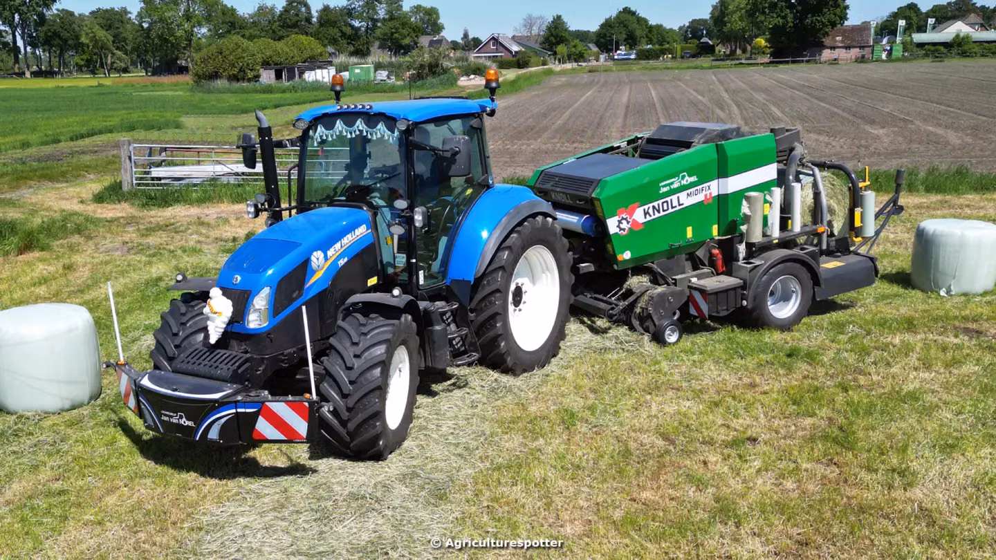 A blue tractor with a black front weight bumper works in a grassy field towing green baling equipment