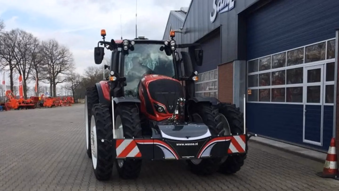 A red tractor fitted with a front weight bumper featuring reflective stripes and a website URL, parked outside a building
