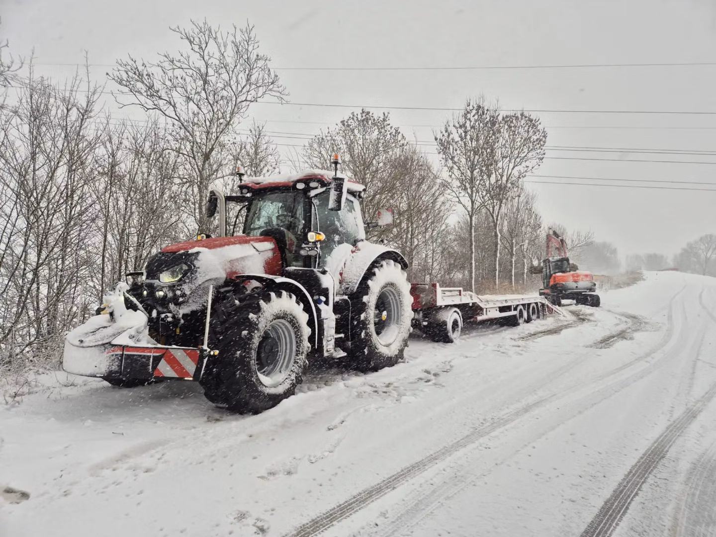 Tractor with a front weight bumper and snow-covered tires is parked on a snowy road with a trailer and excavator behind
