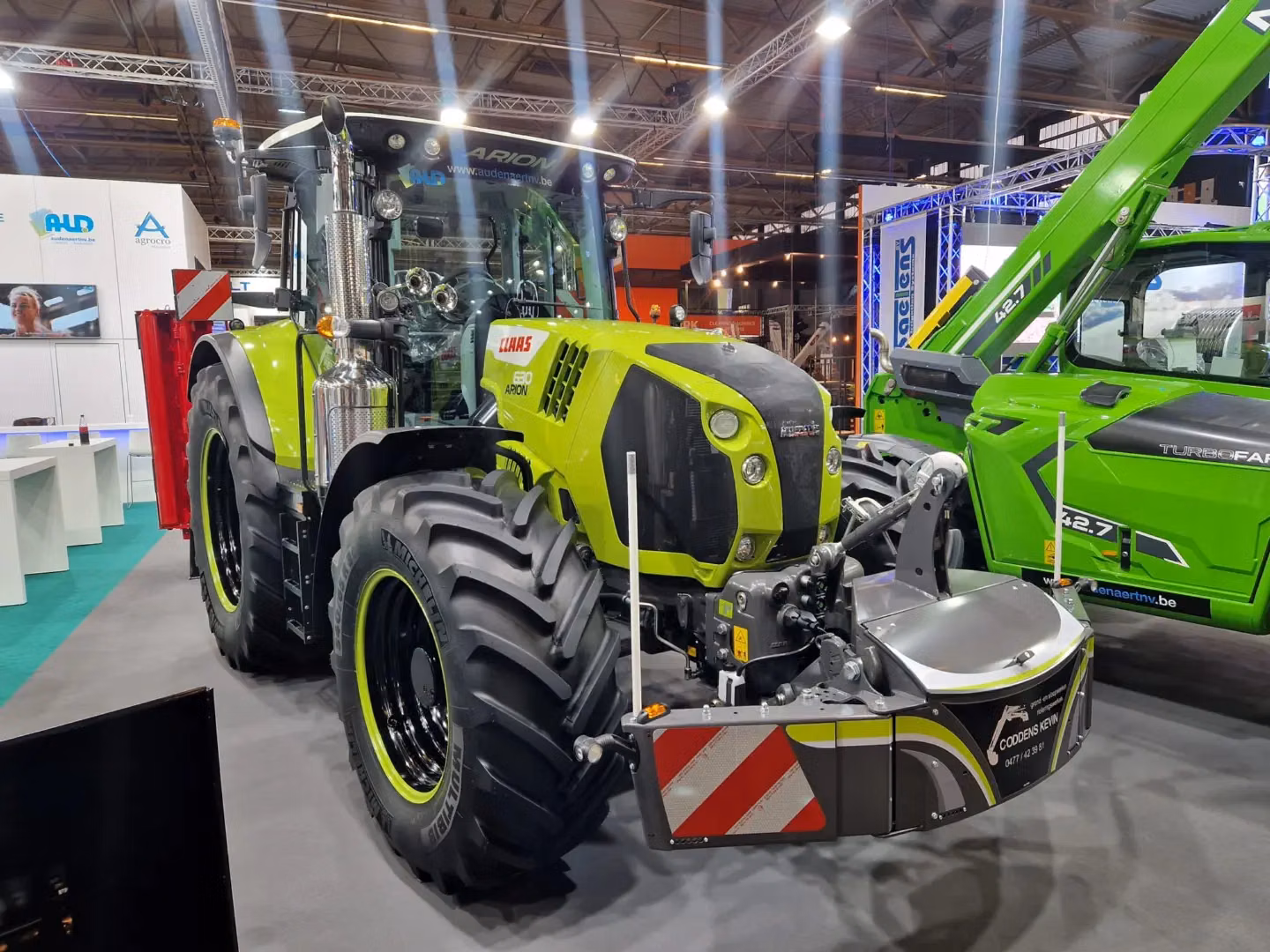 A green tractor on display indoors with a large front weight bumper attached and red and white safety stripes visible