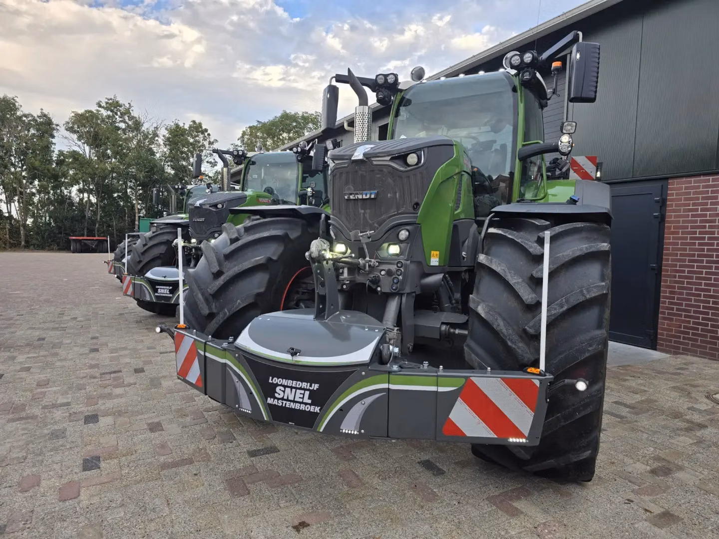 Two green tractors parked outside a building, each fitted with a large front weight bumper with red and white safety stripes
