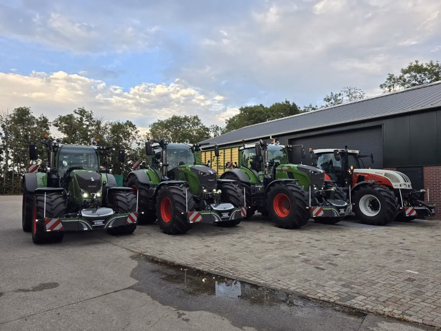 Four tractors equipped with large front weight bumpers are parked side by side on a paved lot near a building